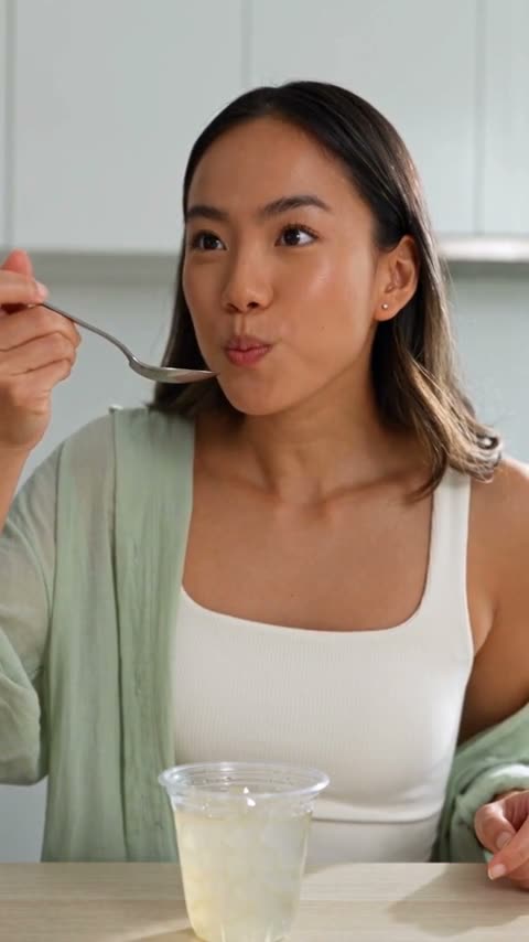 A woman peels open a cup of aloe vera cubes, lifts a spoonful of the clear jelly, tastes it and smiles, with the tub set beside a bunch of grapes.
