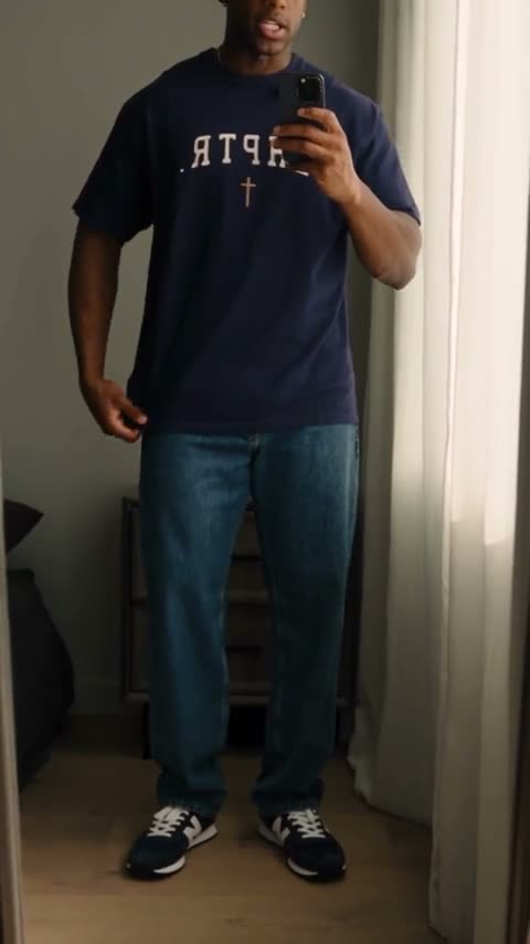 A man films a mirror selfie in a navy CHPTR T-shirt, tugs at the heavyweight cotton, steps back for the full fit with jeans and trainers, then adds a cap.