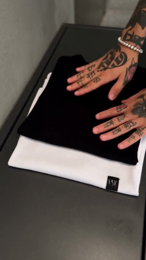A tattooed man in a black slim-fit tee climbs a concrete stairwell, carries in a delivery box and folds the black and white pair on a counter.