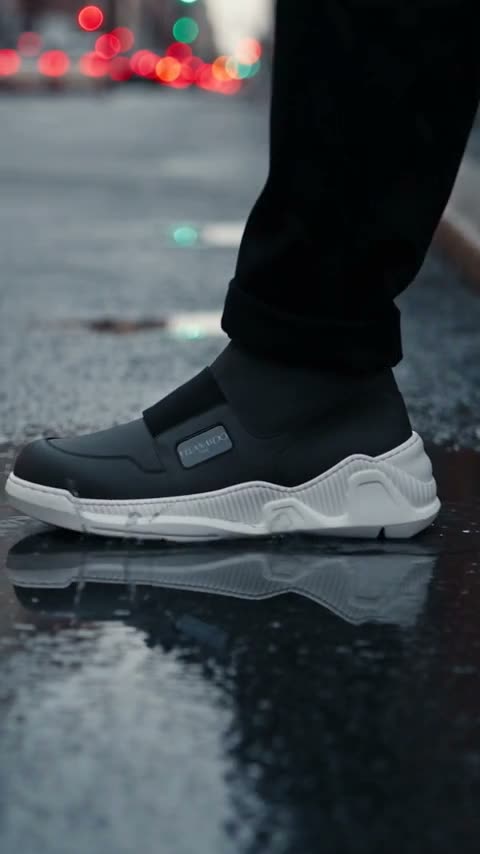 Black strap-front boots worn across a wet city street: ankle-height tracking through the puddles on a zebra crossing, the patterned outsole turned to camera on a kerb step, then a side profile walking past a stone wall.